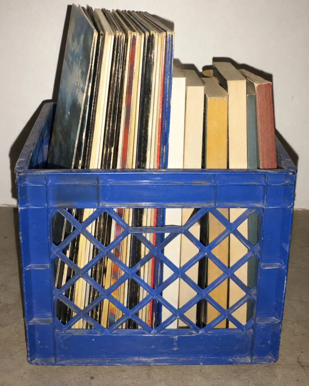 A blue plastic milk crate packed upright with records and paperbacks, lit by a hard flash